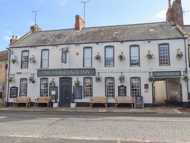 The front facade of The Hermitage Inn with benches and hanging flower baskets at Dray Cottage in Warkworth