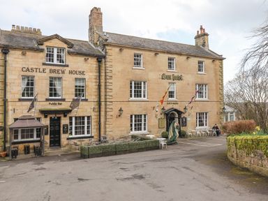 A stone building with signs for Castle Brew House and Sun hotel with outdoor seating and flags at Dray Cottage in Warkworth