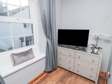 A corner of a room with a white window seat with a cushion gray curtains a gray dresser with a flat screen TV and a white flower at Dray Cottage in Warkworth