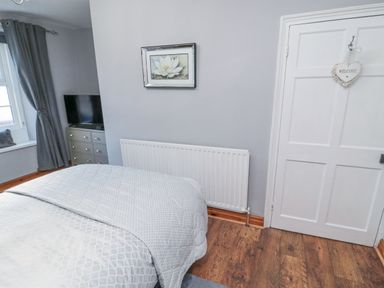 A bedroom with a bed covered in patterned bedding a white radiator on the wall a dresser with a television and a window with gray curtains at Dray Cottage in Warkworth