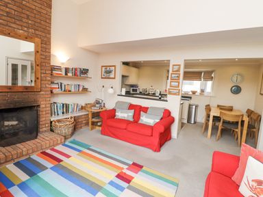 A living room with a brick fireplace red sofas a multicolored striped rug bookshelf and a dining area with a wooden table and chairs at Dunes Cottage in Beadnell