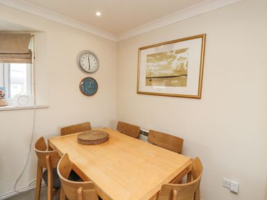 A dining area with a wooden table and six chairs a clock and barometer on the wall a framed picture and a small window with a blind at Dunes Cottage Beadnell