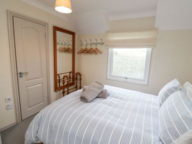 A bedroom with a bed covered with striped bedding two rolled towels on the bed a window with a blind a door a large mirror and a rack with hangers at Dunes Cottage in Beadnell