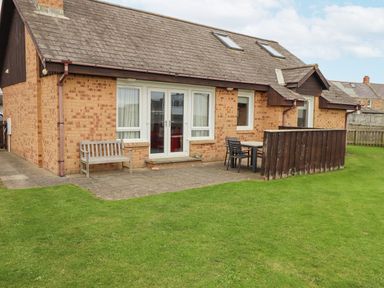 A brick house with a tiled roof patio with wooden bench and table with chairs on grass at Dunes Cottage Beadnell