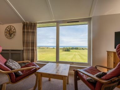 A sitting area with two armchairs and a wooden table by a large window overlooking grassy land and the sea at Fidra in Embleton