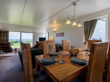 A dining area with wooden table and chairs set with plates and glasses next to a living room with chairs and a TV at Fidra in Embleton