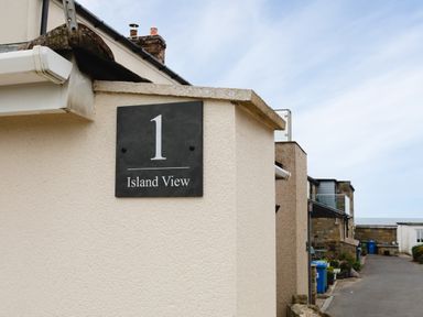 A street view showing a wall with a sign reading 1 Island View and buildings alongside a road at Island View in Amble