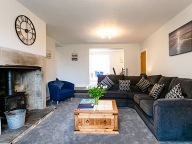 A living room with a gray sectional sofa and patterned cushions a wooden coffee table with flowers a blue armchair a fireplace with a wood stove and a large wall clock at Island View in Amble
