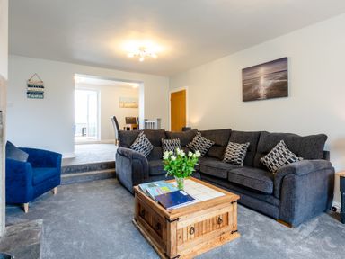 A living room with a dark gray sectional sofa with pillows a wooden coffee table with books and flowers a blue armchair and an open doorway leading to a dining area at Island View in Amble