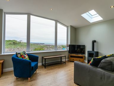A sitting room with a blue armchair a gray sofa a wooden TV stand with a TV and a small wood stove by large windows at Island View in Amble