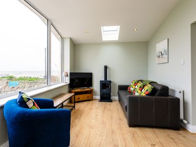 A living room with a blue armchair a black sofa colorful cushions a small wood stove a tv on a wooden stand and large windows at Island View in Amble