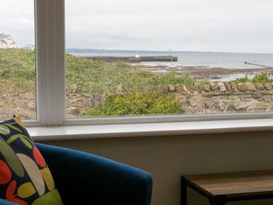 A window view of a stone wall bushes and a sea with a pier seen from inside a room with a blue chair and colorful cushion at Island View in Amble