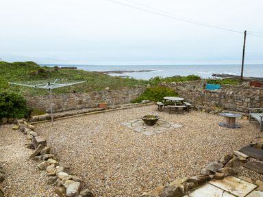A gravel courtyard with a rotary clothesline a round picnic table a fire pit and stone walls overlooking the sea at Island View in Amble