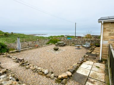 An outdoor gravel patio area with a round wooden picnic table a bench a small round table and a stone wall overlooking the sea at Island View in Amble
