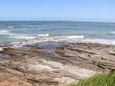 Rocky shoreline with waves and a small island with a lighthouse in the distance at Island View in Amble
