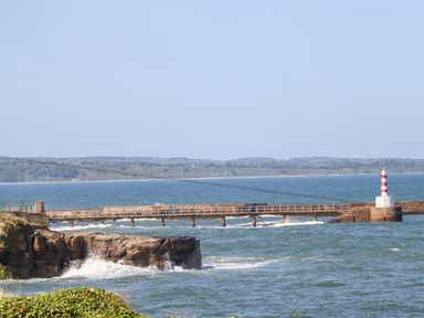 A pier with a red and white striped lighthouse extending over the sea with waves crashing at rocky cliffs and land visible in the background at Island View in Amble