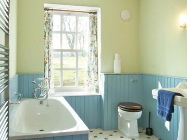 A bathroom with a bathtub, toilet with dark seat, sink with blue towel, window with floral curtains, and blue wainscoting at Kirknowe in Jedburgh
