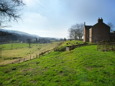 A stone house on a grassy hill with fenced areas and rolling countryside in the background at Kirknowe in Jedburgh