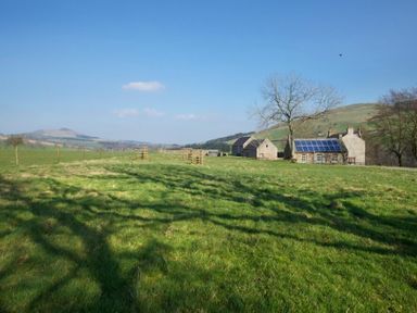 A grassy field with a fenced area and stone buildings with solar panels in the background at Kirknowe Jedburgh