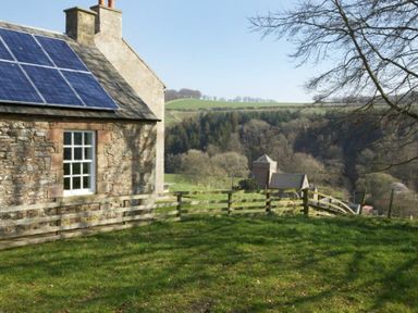 A stone house with solar panels on the roof and a wooden fence in a grassy yard overlooking trees and hills at Kirknowe in Jedburgh