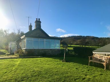 A stone house with a glass sunroom surrounded by a lawn and countryside at Leazes Cottage in Rothbury
