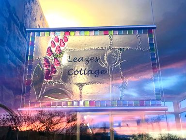 A glass sign with flowers and text Leazes Cottage with a reflection of trees and sunset in a window at Leazes Cottage in Rothbury