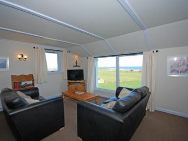 A living room with two black sofas a wooden coffee table a TV on a wooden stand and large windows overlooking a field at Longstone in Embleton