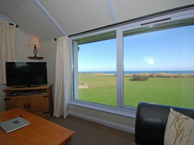 A living room with a television on a wooden cabinet a coffee table a black sofa and large windows showing a green field and sea at Longstone in Embleton