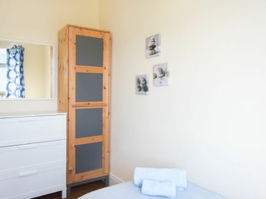 A bedroom corner with a white dresser, a wardrobe with grey panels, a mirror, three pictures on the wall, and folded towels on a bed at Melvin Cottage in Low Newton-By-The-Sea