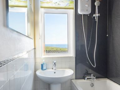A bathroom with a sink and soap dispenser next to a bathtub and shower with a window showing a field and sea view at Melvin Cottage in Low Newton-By-The-Sea