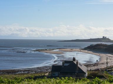 A coastal scene with a house near a sandy beach and a castle ruin on a hill in the distance at Melvin Cottage in Low Newton-By-The-Sea