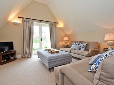 A living room with beige sofas, a large ottoman, lamps, a TV on a wooden stand, and doors with curtains at Sandpiper Bamburgh