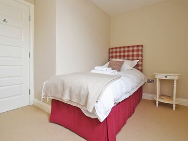 A bedroom with a single bed with red bed skirt and checked headboard and a small round bedside table with books at Sandpiper Bamburgh