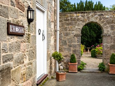 A stone building entrance with a white door and a sign reading Mid Cote with an arched stone gateway leading to a garden with potted plants at Mid Cote Shoreston near Bamburgh