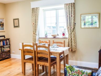 A dining area with a wooden table set for four near a window with patterned curtains and a bookshelf in the corner at Mid Cote Shoreston near Bamburgh