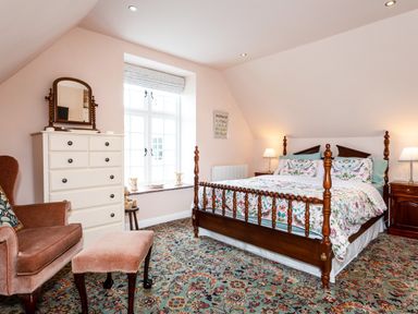 A bedroom with a wooden bed with floral bedding a white dresser with a mirror a pink armchair and ottoman and a floral carpet at Mid Cote Shoreston near Bamburgh