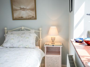 A bedroom with a metal bed, floral bedding, a wicker nightstand, a table lamp, and a window with bowls on the sill at Mid Cote Shoreston near Bamburgh