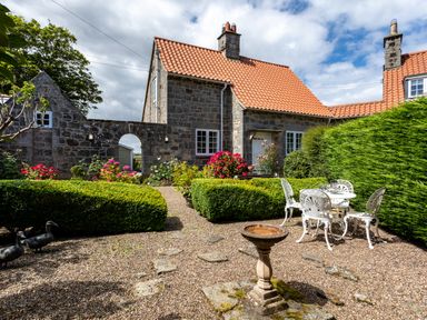 A stone house with a red tile roof and garden with hedges flowers and outdoor white metal table and chairs at Mid Cote in Shoreston near Bamburgh