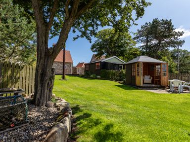 A garden with green grass trees a wooden fence two sheds a stone house and outdoor chairs at Mid Cote Shoreston near Bamburgh