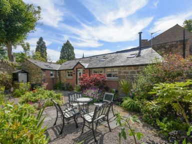 A garden patio with table and chairs surrounded by plants and a stone and brick house in the background at Nurses Cottage in Alnwick