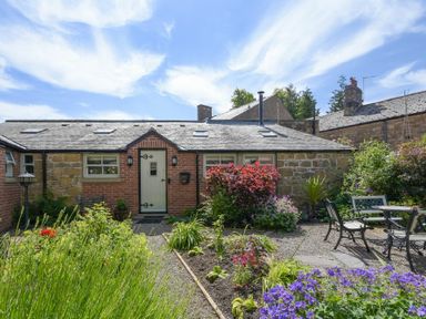 A garden with flowers and outdoor furniture in front of a brick and stone house at Nurses Cottage in Alnwick