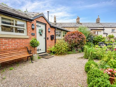 A brick exterior of a cottage with a wooden bench and a gravel path surrounded by green plants and bushes at Nurse's Cottage in Alnwick