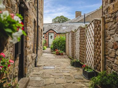 A narrow stone pathway between buildings with flower pots and wooden fencing at Nurse's Cottage in Alnwick