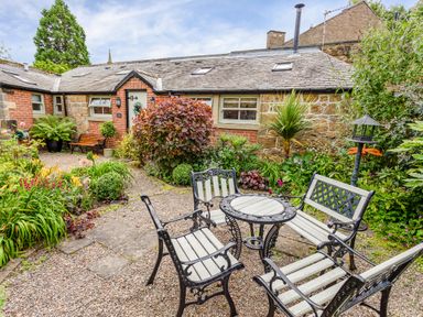 An outdoor garden area with metal benches and a round table surrounded by plants and flowers at Nurses Cottage in Alnwick