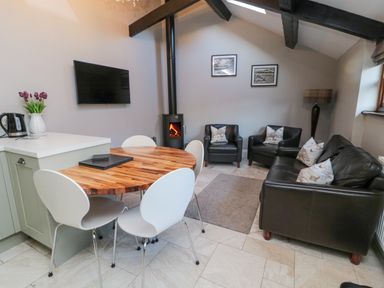 A room with a round wooden dining table and four white chairs a black sofa and two black armchairs with cushions a wood burning stove and a wall mounted TV at Nurse's Cottage in Alnwick