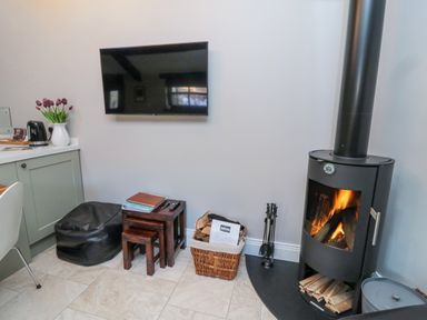 A room corner with a wood burning stove a basket of firewood a black bean bag a stack of nested wooden stools a wall-mounted television and a kitchen counter with a kettle and flowers at Nurses Cottage in Alnwick