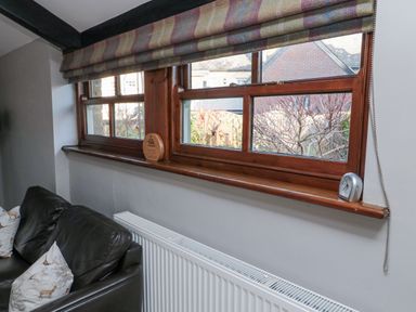 A window with wooden frame and striped curtain above a radiator and next to a black leather chair with animal print cushions at Nurses Cottage in Alnwick