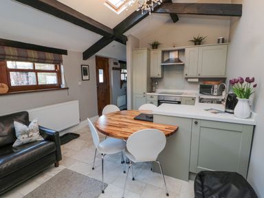 A kitchen and dining area with wooden beams a wooden table white chairs and a black sofa at Nurses Cottage in Alnwick