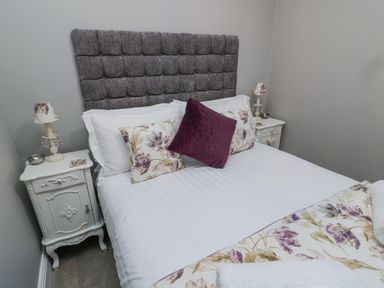 A bedroom with a bed with a gray headboard and floral pillows and two white bedside tables with lamps at Nurse's Cottage in Alnwick