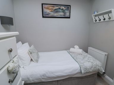 A small bedroom with a single bed white bedding and a floral pillow a chest of drawers a wall-mounted coat rack and a painting above the bed at Nurse's Cottage Alnwick